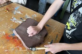 Eine Person bearbeitet mit den Händen ein Stück braunen Ton auf einem Tisch.